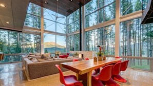 Une pièce spacieuse avec une grande table à manger en bois et des chaises rouges, un canapé et une vue sur une forêt, visible à travers de grandes fenêtres.