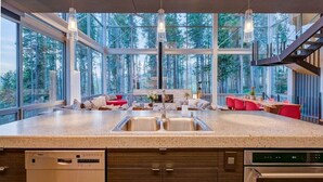 Une cuisine moderne dotée d’un grand îlot central, d’un lave-vaisselle encastré et d’un four. De grandes fenêtres offrent une vue sur la forêt environnante.