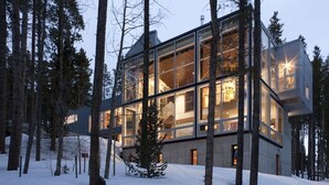Une maison moderne en verre, entourée de grands arbres, se dressant au milieu d’un paysage enneigé.