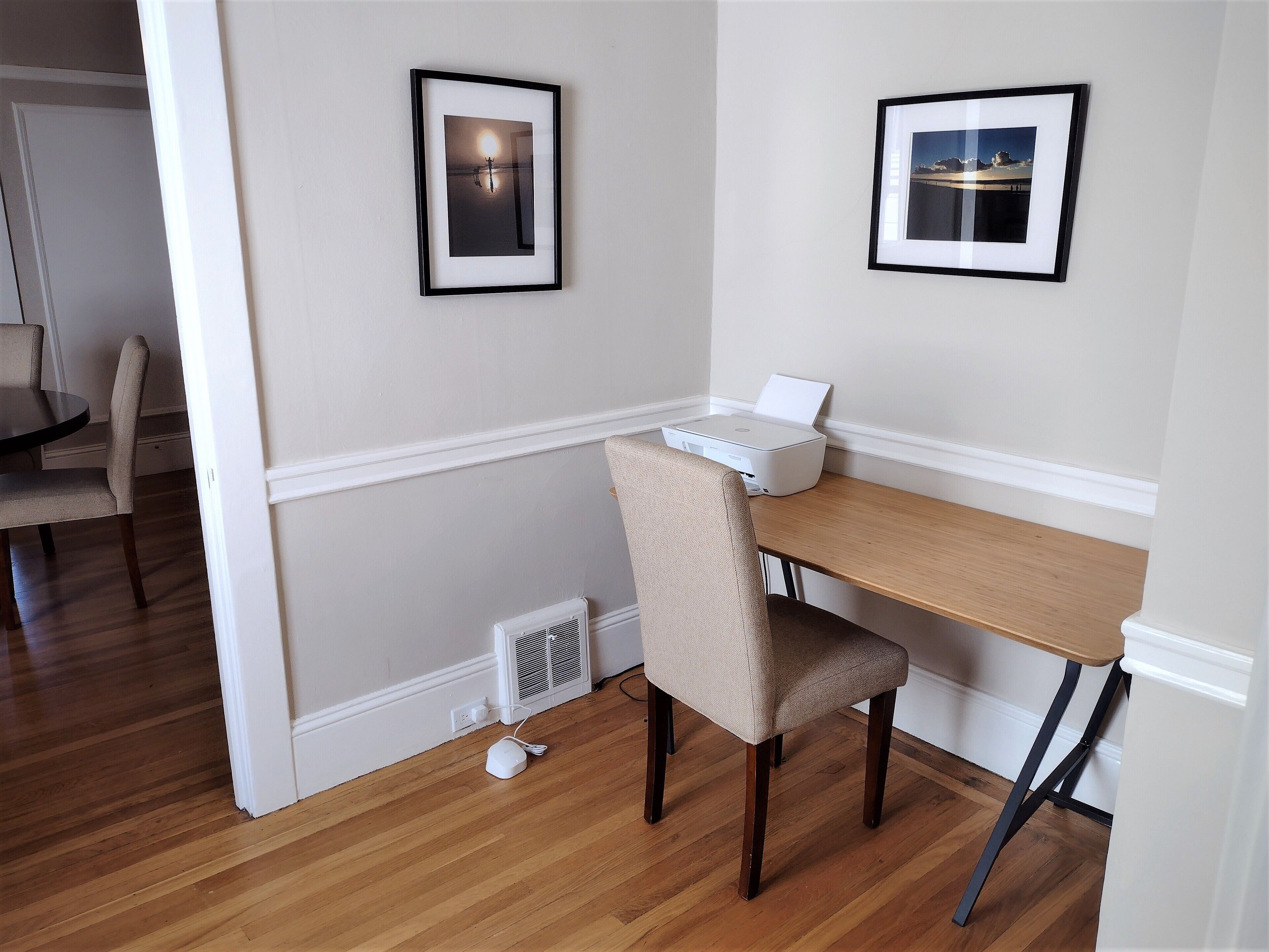 A room with a wooden desk, a chair, a printer, and two framed pictures on the wall.