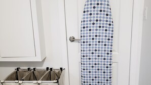 Una puerta blanca con una tabla de planchar azul con lunares. Hay tres cajas de almacenamiento en el suelo, etiquetadas como “LIGHT”, “COLOR” y “DARK”.