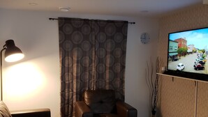 Una sala de estar con un sofá de cuero marrón, un sillón blanco, una mesa de centro, una lámpara, un televisor montado en la pared y una cortina con diseño.