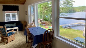 Une pièce chaleureuse avec un plafond en bois, une grande fenêtre donnant sur un lac, une table à manger avec des chaises et un canapé recouvert d’un tissu bleu.