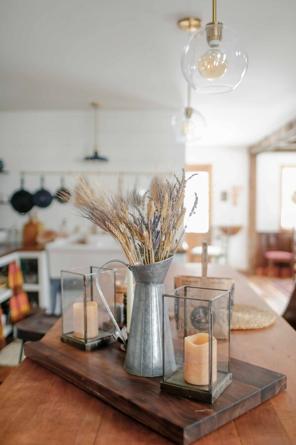 A kitchen counter with a rustic wooden table, a metal pitcher with dried flowers, and two glass candle holders.
