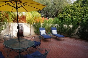 An outdoor patio with a round metal table and blue cushioned chairs, a yellow umbrella, and a brick floor.