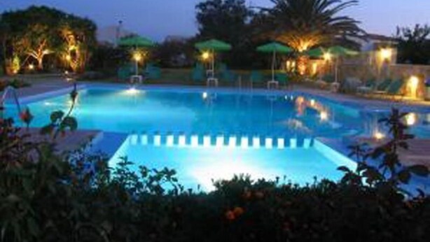 A swimming pool with a clear blue water surface, surrounded by lounge chairs and umbrellas, and illuminated by ambient lighting. - Androulakis Apartments (Platanias)