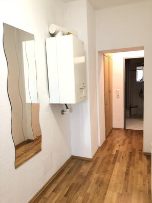 A hallway with wooden flooring, a white wall with a mirror and a white cabinet, and a wooden door leading to another room.