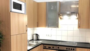 A compact kitchen with wooden cabinets, a microwave, a kettle, a stove, and a dishwasher. - Wildbach City Apartments (Zell am See)