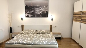 A bedroom with a bed, bedside tables, a nightstand, a wall-mounted picture, and a wooden floor. - Wildbach City Apartments (Zell am See)