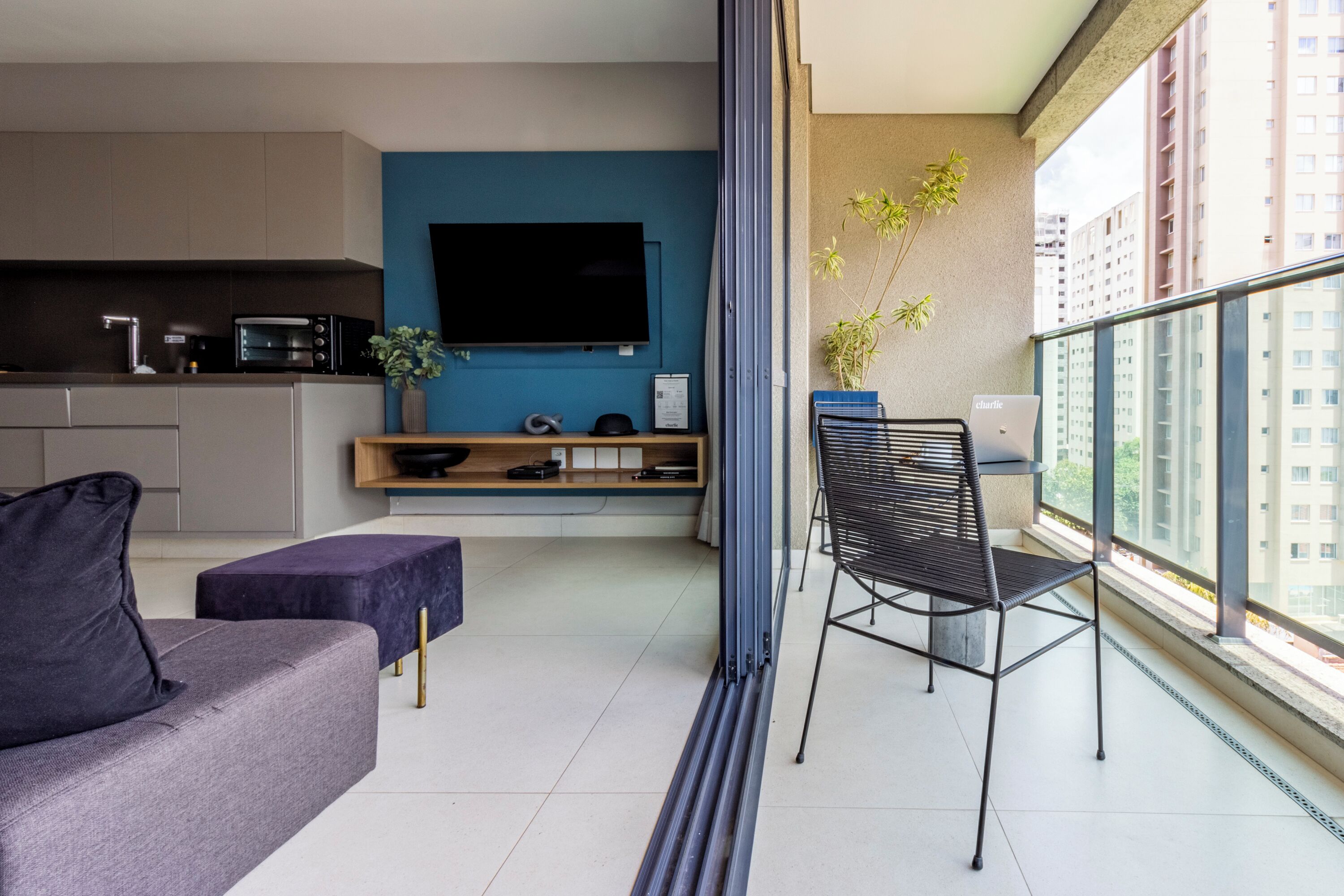 A modern living room with a sofa, a dining table, and a TV mounted on the wall. There is a balcony with a chair and a plant.