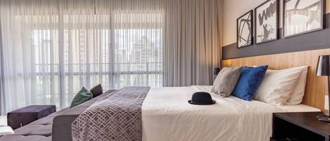 Quarto de hotel moderno com uma cama grande, uma mesa de cabeceira e uma janela com cortinas translúcidas.