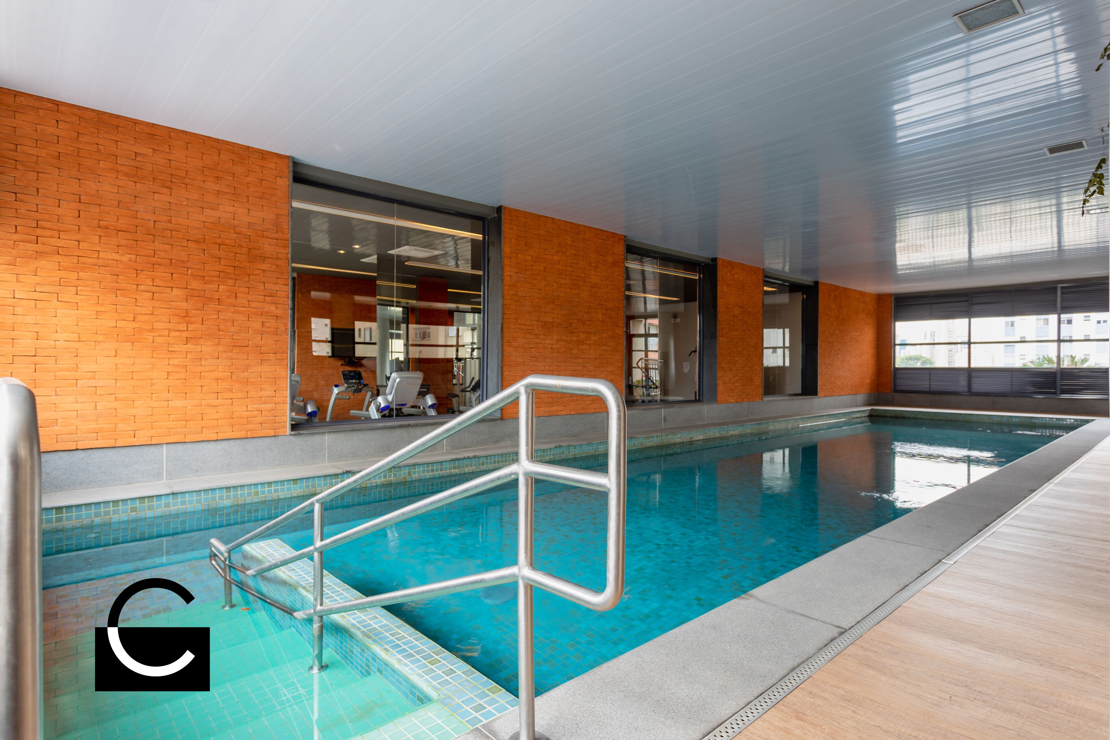An indoor swimming pool with a clear view of the surrounding area, including a brick wall and a glass door leading to a room with exercise equipment.