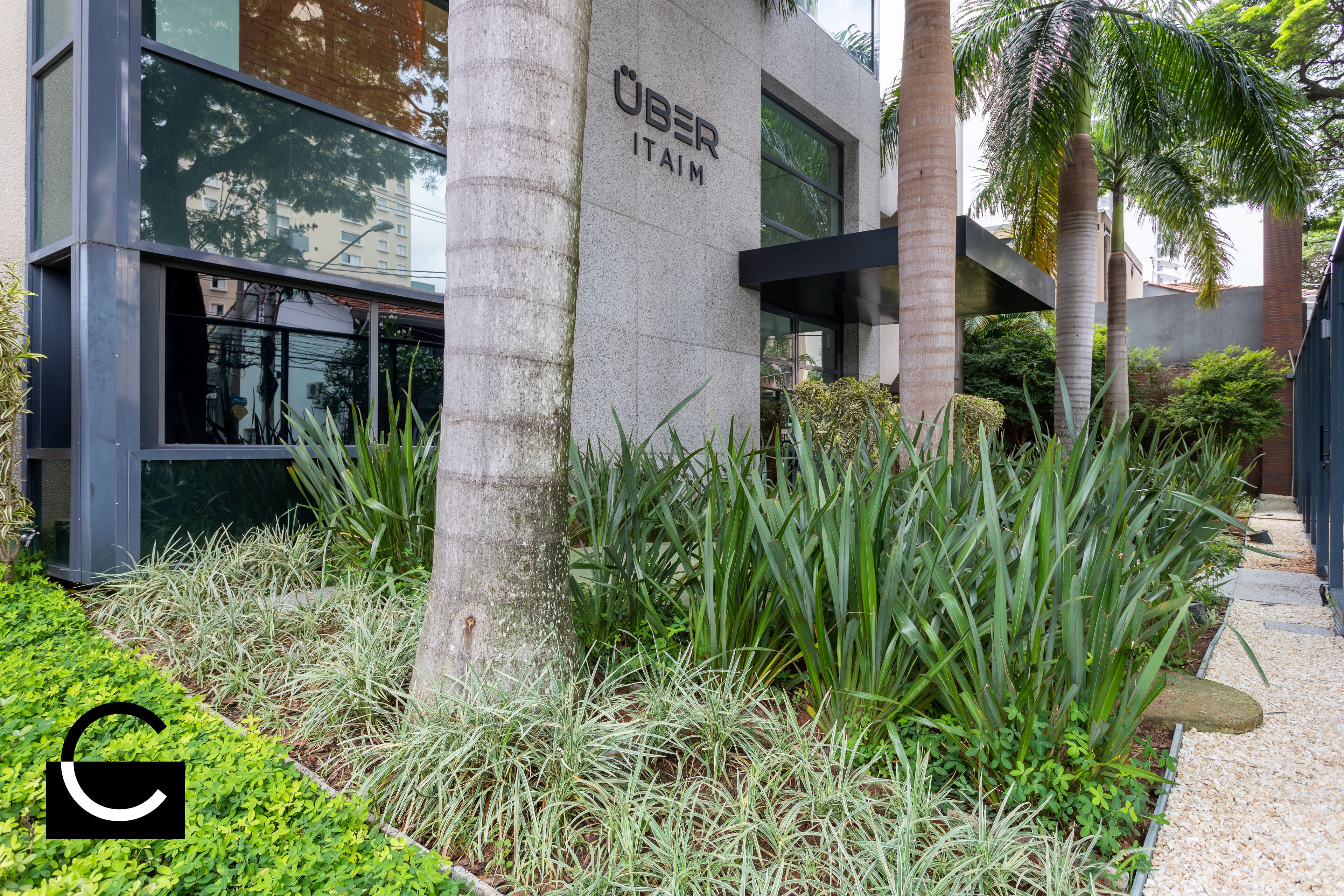 Un edificio moderno con el letrero "UBER ITAIM" en su fachada, rodeado de exuberante follaje y palmeras.