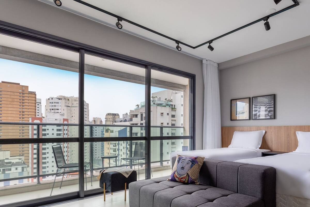 A modern hotel room with large windows, a sofa, two beds, and a view of the cityscape.