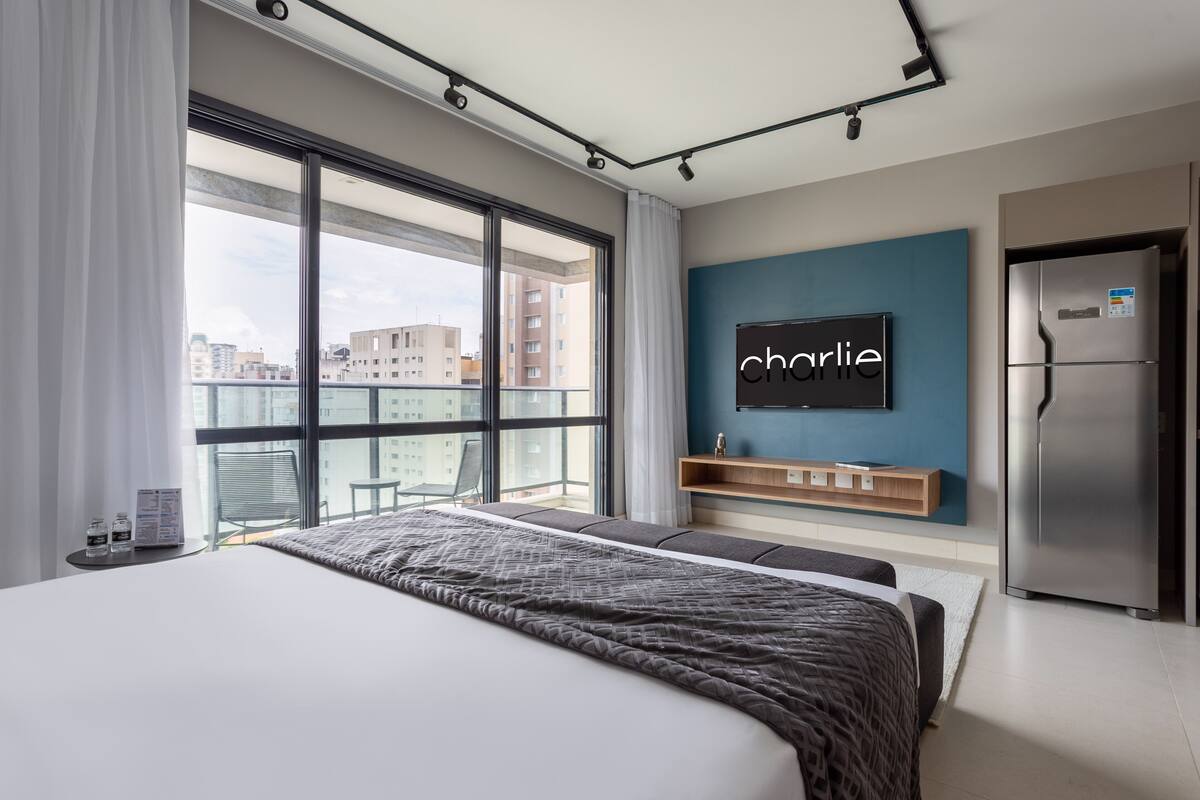 A modern hotel room with a large bed, a TV mounted on the wall, a refrigerator, and a balcony with a city view.