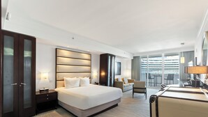 A modern hotel room with a large bed, a desk with a lamp, a wardrobe, and a view of the city through large windows.