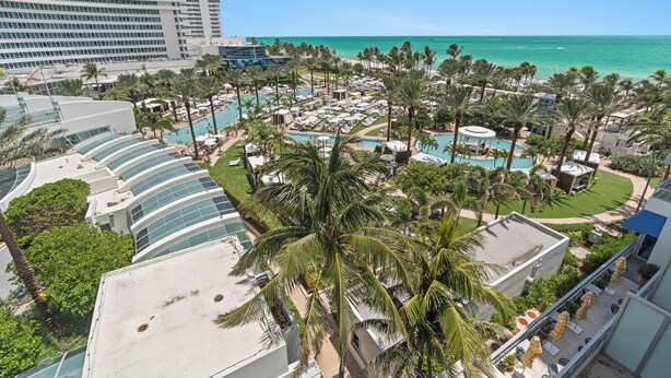A resort with a large pool surrounded by palm trees and lounge chairs, a clear view of the ocean, and a modern building in the background.