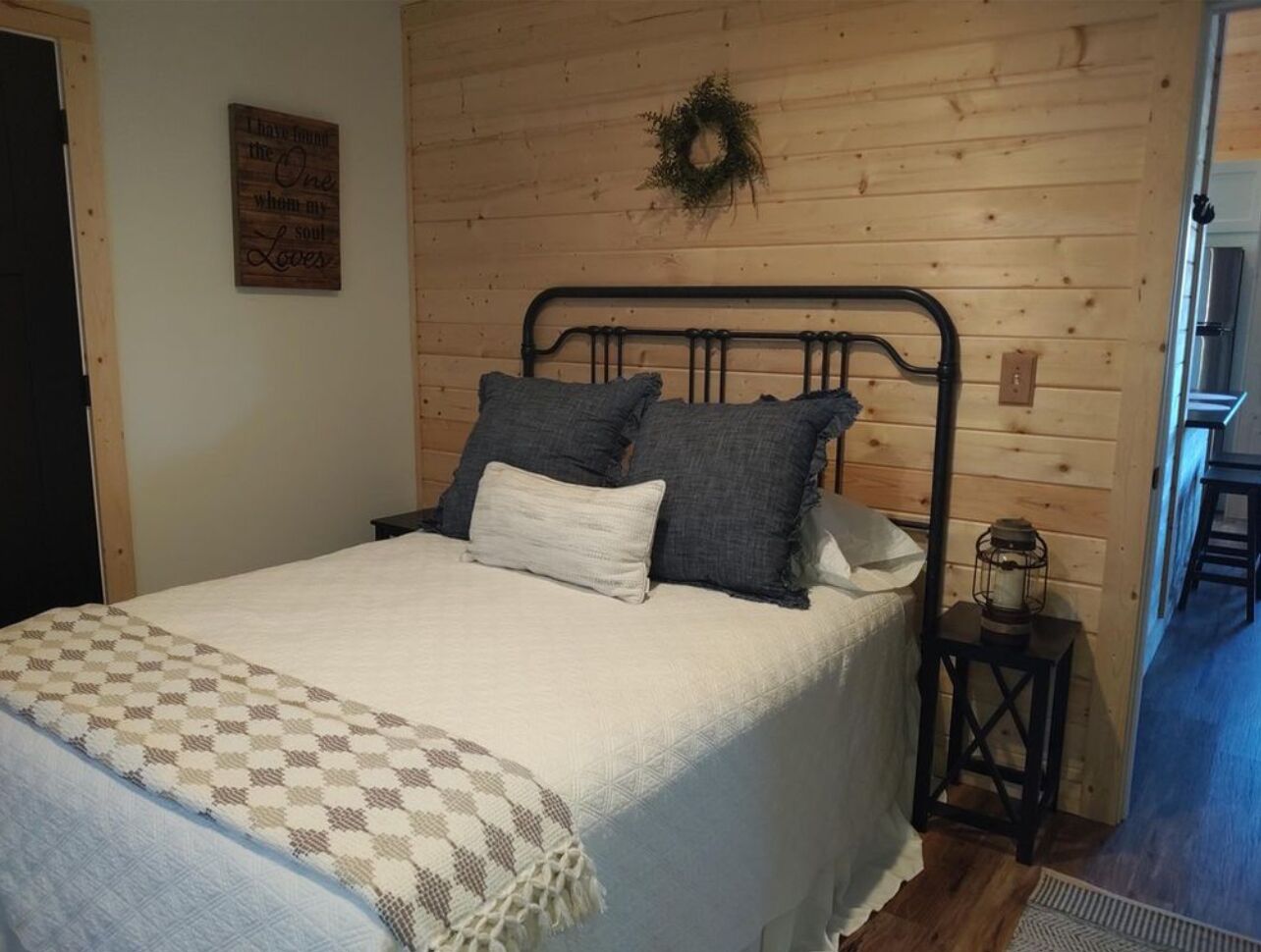 A bedroom with a bed, a wall-mounted wreath, a small table with a lantern, and a checkered blanket.