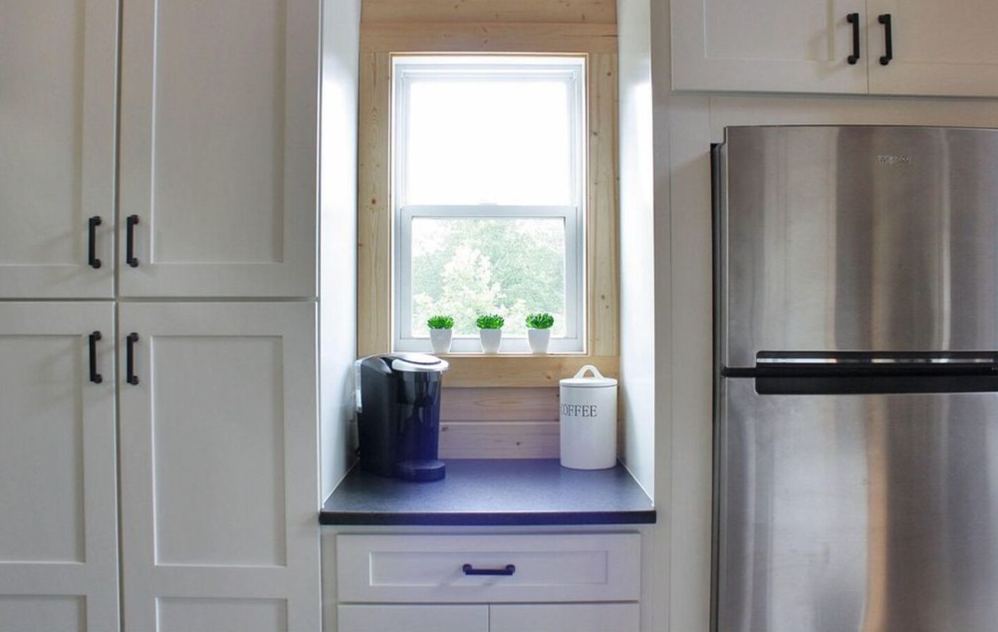A kitchen with white cabinets, a stainless steel refrigerator, a window with potted plants, and a coffee maker.
