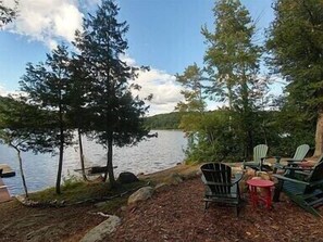 A lakeside seating area with Adirondack chairs and a small table, surrounded by trees and a dock.