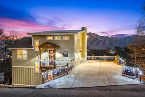 A two-story house with a porch and a covered front entrance, surrounded by mountains and decorated with string lights.