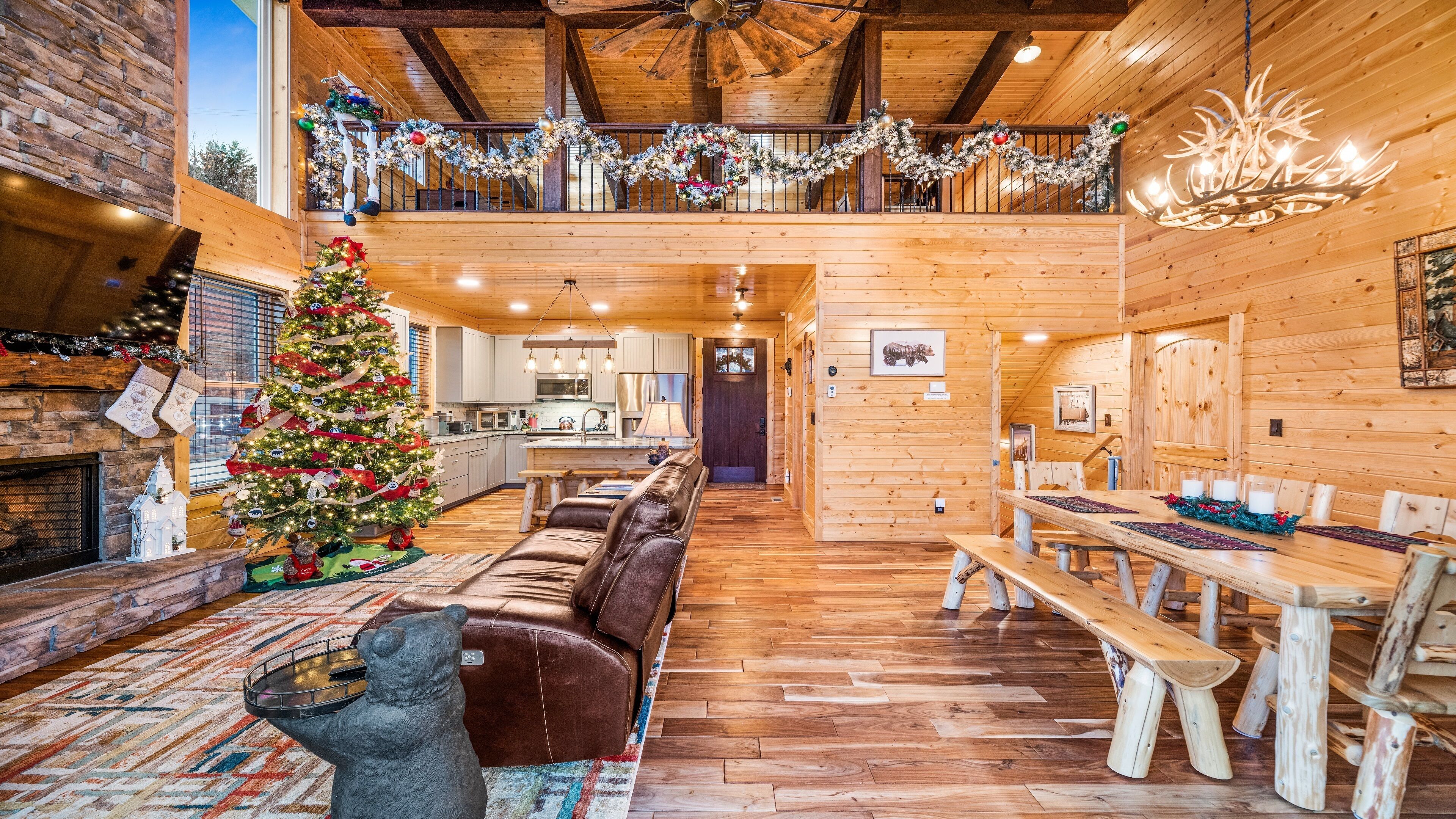 A cozy log cabin interior with a wooden dining area, a fireplace, a Christmas tree, and a loft with a balcony.
