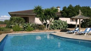 A swimming pool with lounge chairs, surrounded by palm trees and a building with a tiled roof. - Villa Llobets I (Llucmajor)