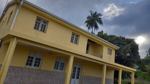 A two-story building with a yellow facade, multiple windows, and a stone base. - Apartement F3 en bas de Villa (Basse-Pointe)