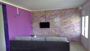 A room with a purple wall, a gray sofa, a mounted TV, and a window. - Apartement F3 en bas de Villa (Basse-Pointe)