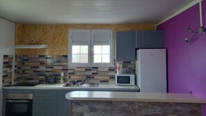 A kitchen with a stone backsplash, a microwave, a refrigerator, and a window with shutters. - Apartement F3 en bas de Villa (Basse-Pointe)
