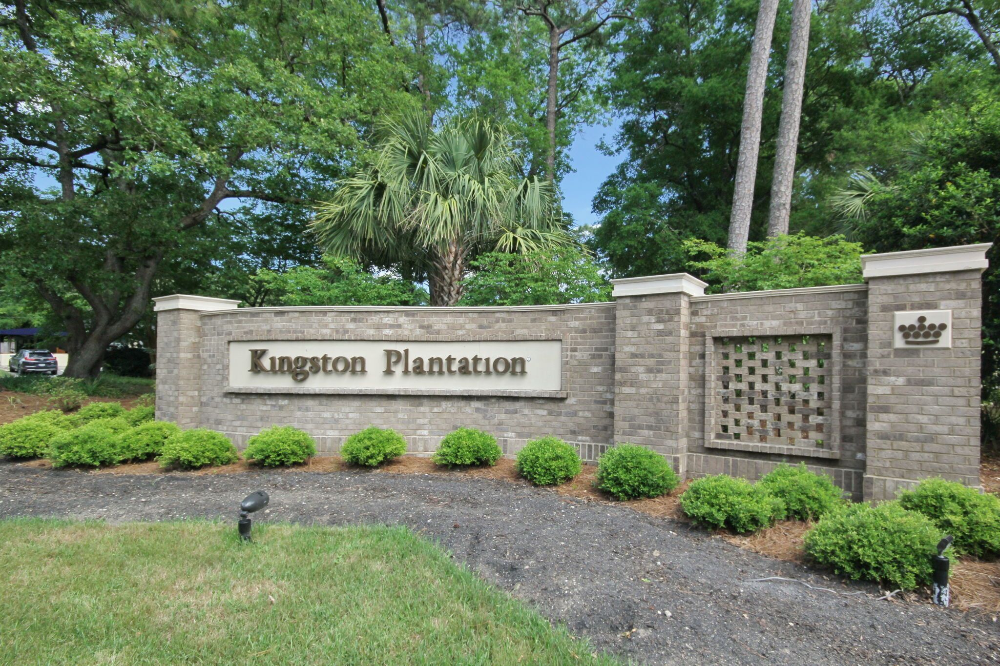 A sign for Kingston Plantation with a brick wall, a palm tree, and neatly trimmed bushes.