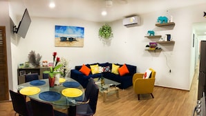 Un salon comprenant une table à manger en verre, des chaises bleues et jaunes, un canapé bleu foncé avec des coussins orange, un fauteuil jaune et une table basse.