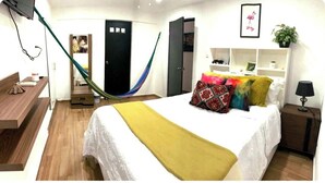 Une chambre d’hôtel comprenant un lit, un bureau, une chaise, un hamac, une armoire et une étagère avec des objets décoratifs.