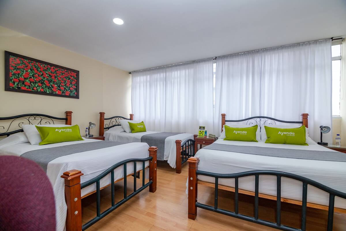 Ein Hotelzimmer mit drei Betten, jeweils mit Holzrahmen und weißer Bettwäsche. Grüne Kissen mit dem Namen des Hotels sind darauf. An der Wand hängt ein Gemälde mit roten Blumen, und ein Fenster mit weißen Vorhängen ist zu sehen.
