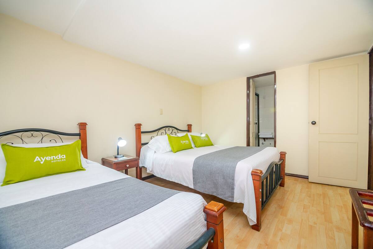 Ein Hotelzimmer mit zwei Betten, jeweils mit grauer Bettdecke und einem grünen Kissen mit dem Namen des Hotels. Ein Nachttisch mit Lampe, eine Holztür und ein Spiegel an der Wand sind ebenfalls vorhanden.