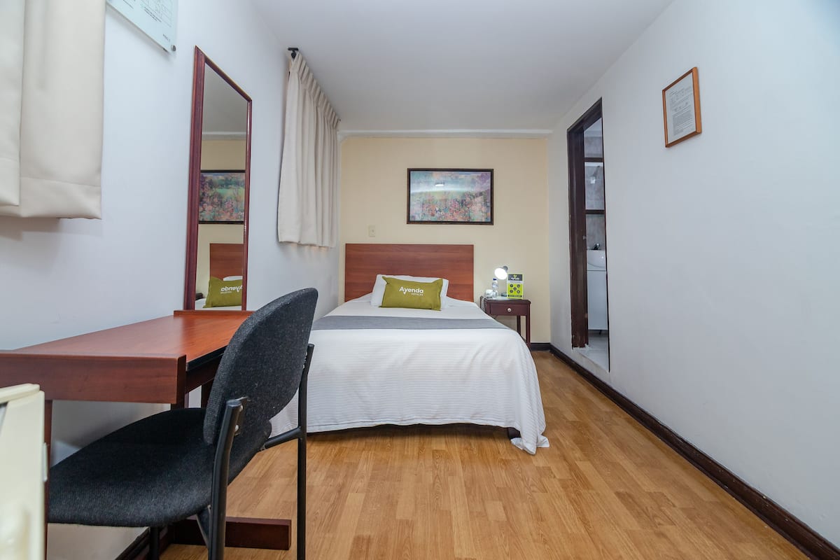 Ein Hotelzimmer mit einem Holztisch, einem schwarzen Stuhl, einem Bett mit weißer Bettdecke und einem grünen Kissen, einem Nachttisch mit Lampe und einem Bild an der Wand.