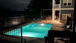 Une piscine rectangulaire, bordée d’une terrasse carrelée et entourée d’une clôture en métal. Un bâtiment à deux étages, doté de balcons et d’un patio couvert, est situé à côté de la piscine.