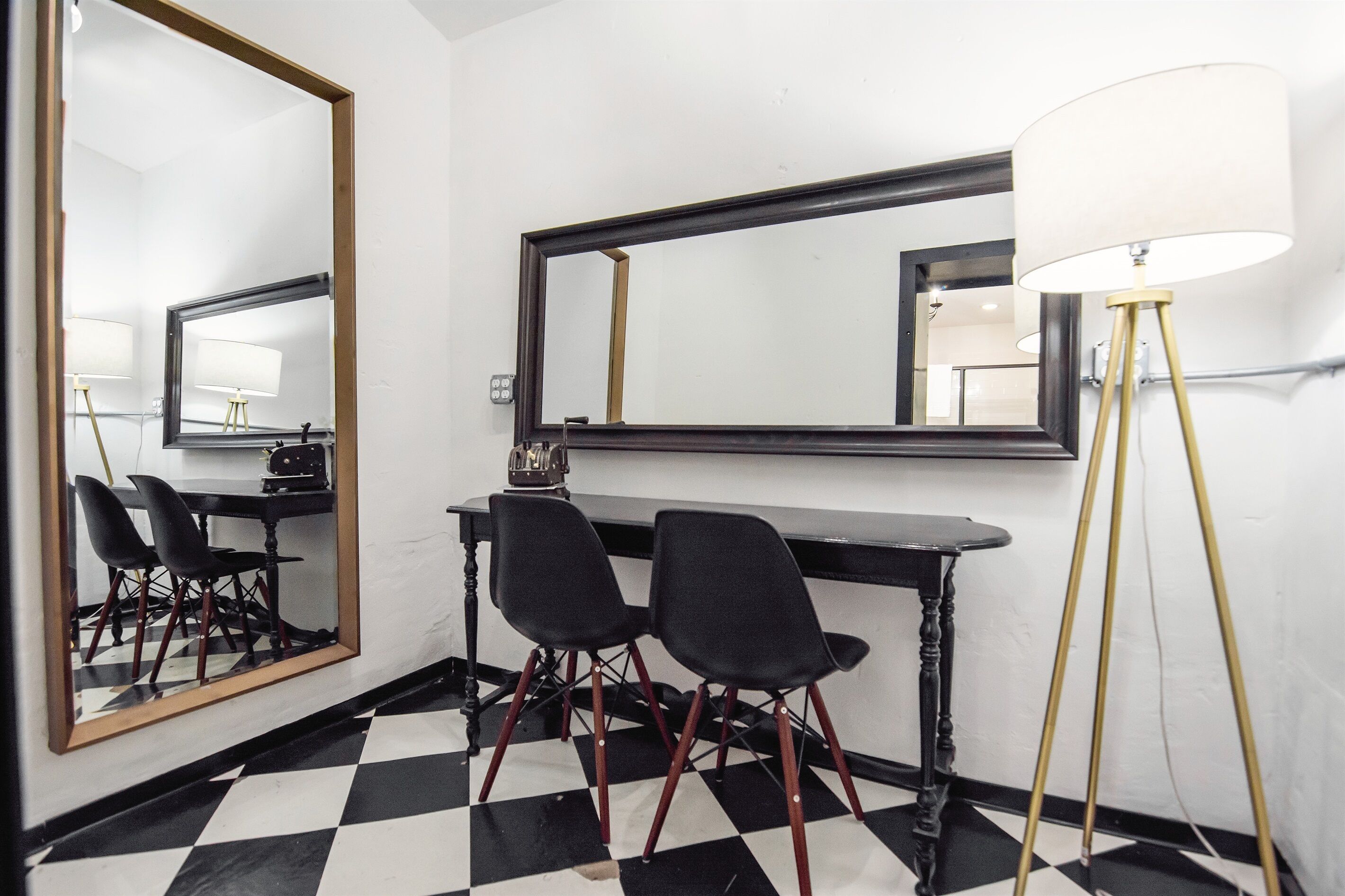 Une pièce avec un sol à carreaux noirs et blancs, un bureau noir avec deux chaises noires, un grand miroir et un lampadaire.