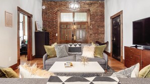 Ein Wohnzimmer mit Backsteinwand, Holztür, Fernseher und einer Couch mit gemusterten Kissen.
