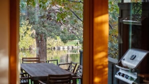 Blick aus einem Zimmer mit Holztisch und -stühlen, einem Grill und einem Teich mit Bäumen und einem Zaun im Hintergrund.