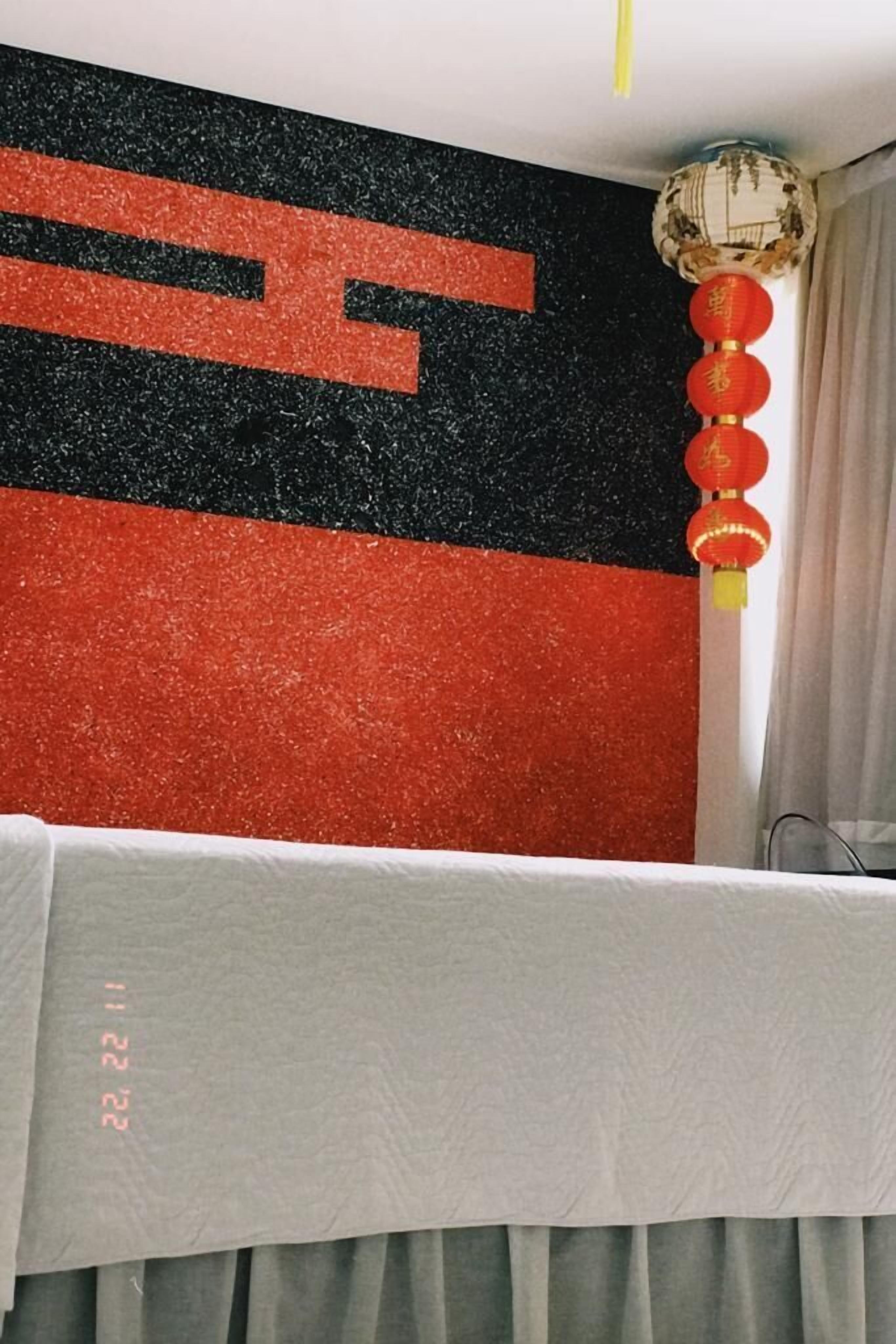 Ein Zimmer mit einer Wand mit Struktur, die ein großes rotes und schwarzes Design aufweist, ein weißes Bett mit grauem Kopfteil und ein traditioneller roter Laternenhänger am Fenster.