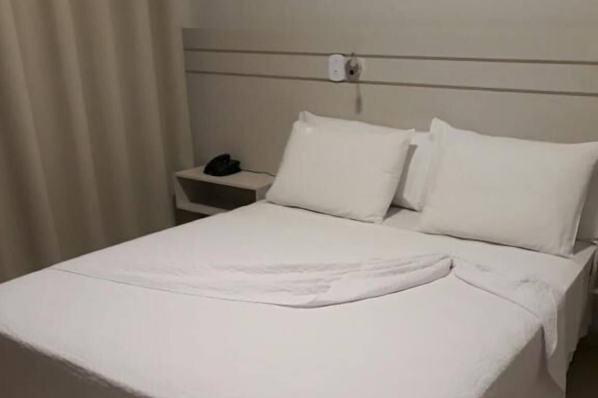 Een opgemaakt bed met witte lakens, een nachtkastje met een telefoon en een lichtschakelaar aan de muur.