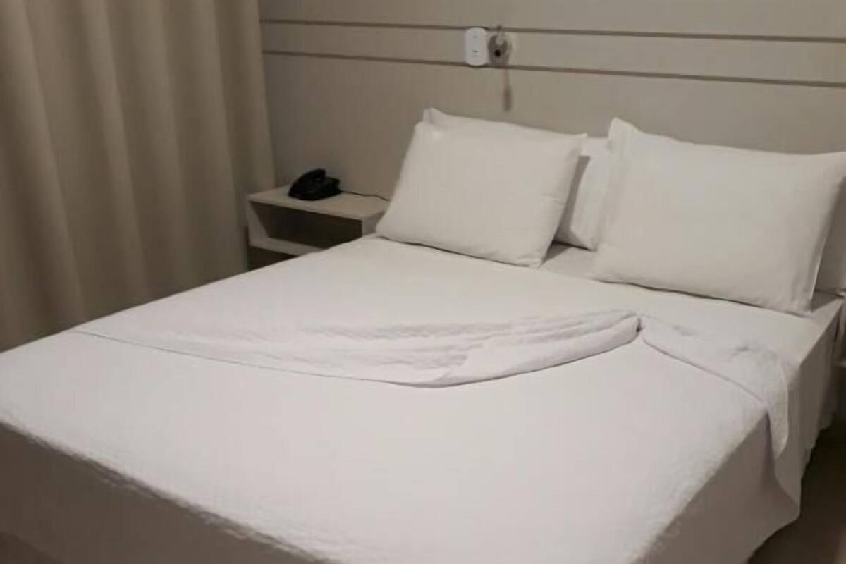Een net opgemaakt bed met witte lakens, een nachtkastje met een telefoon en een stopcontact.