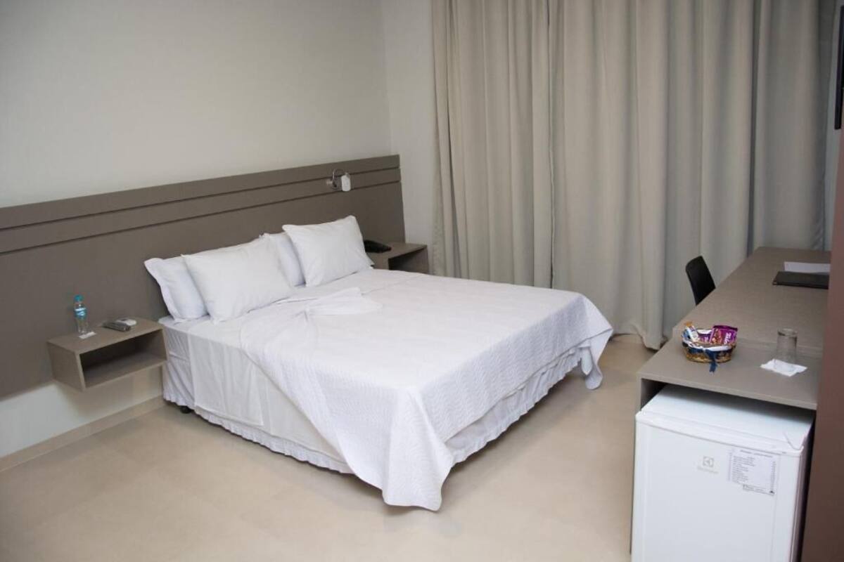 Een hotelkamer met een bed, een bureau, een stoel, een nachtkastje, een lamp, een waterfles en een koelkast.