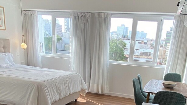 A modern hotel room with a bed, a dining table, and a view of the city through large windows. - Bright & Modern Studio Next to El Olivar Park – San Isidro (San Isidro)