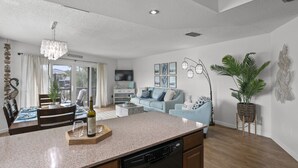 A modern kitchen with a dishwasher, dining table, and chairs. There is a living area with a sofa and a TV.