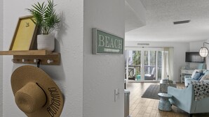 A room with a wooden shelf holding a hat and a framed picture, a potted plant, and a 'BEACH' sign on the wall.
