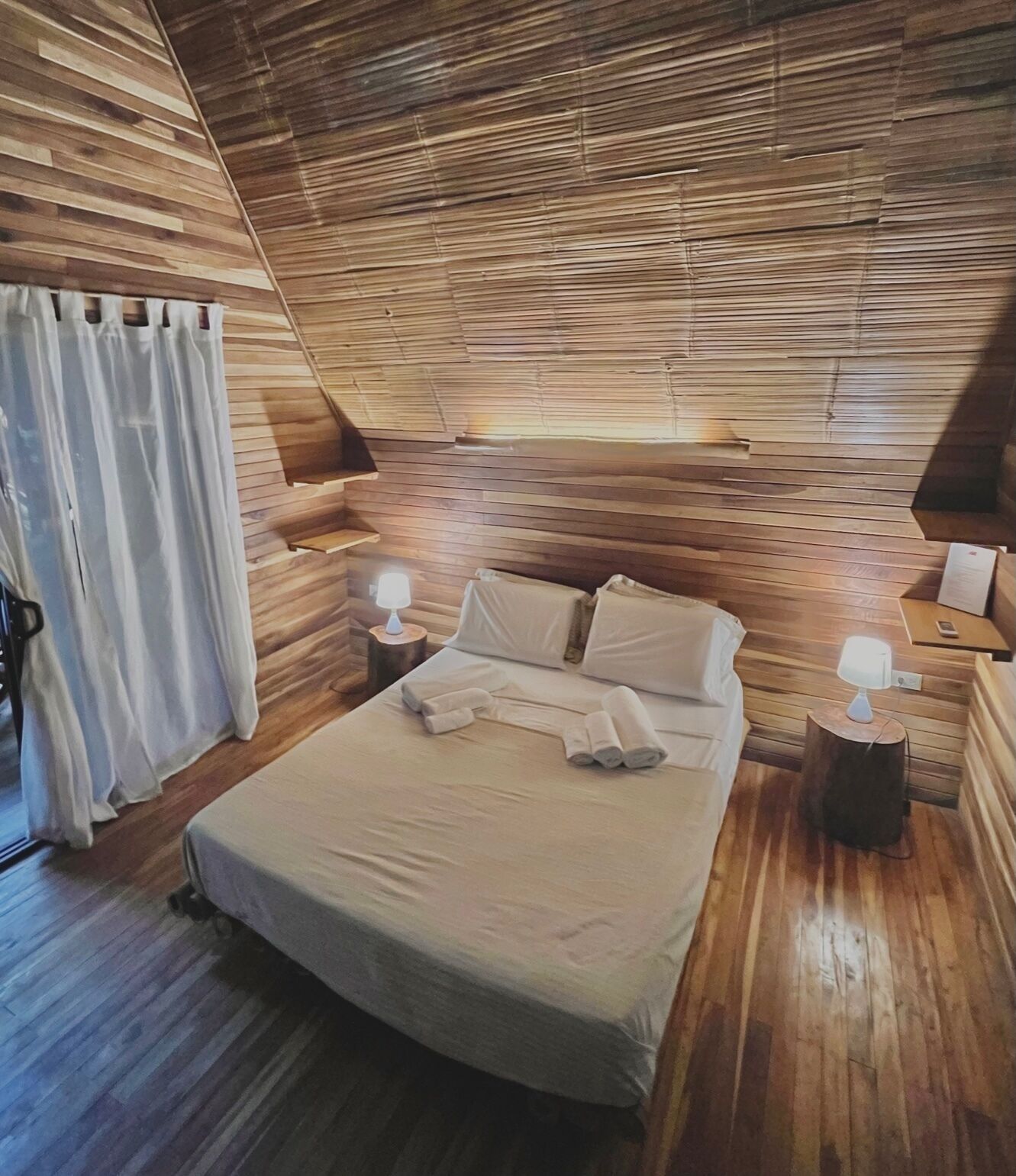 Een slaapkamer met een houten schuine daklijn, een bed met witte lakens, twee bedlampjes en een gordijn aan de linkerkant.