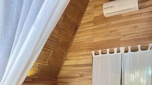 Une chambre avec un plafond en pente en bois, un lit recouvert de draps blancs, une table de chevet avec une lampe et une fenêtre ornée de rideaux légers.