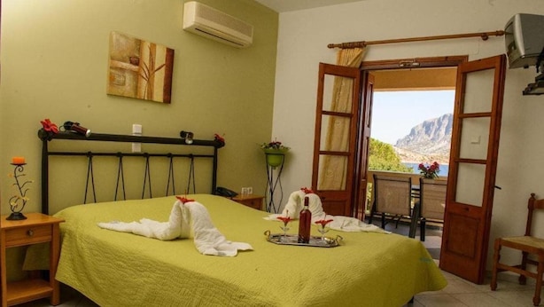 A hotel room with a bed, a bedside table, a window with a view, and a wall-mounted air conditioner. - Amphitrite Apartments (Kalymnos)